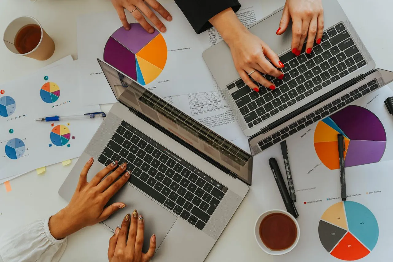 Vista de cima de uma equipe analisando relatórios com gráficos de pizza e de barras coloridos sobre uma mesa, com dois laptops e xícaras de café