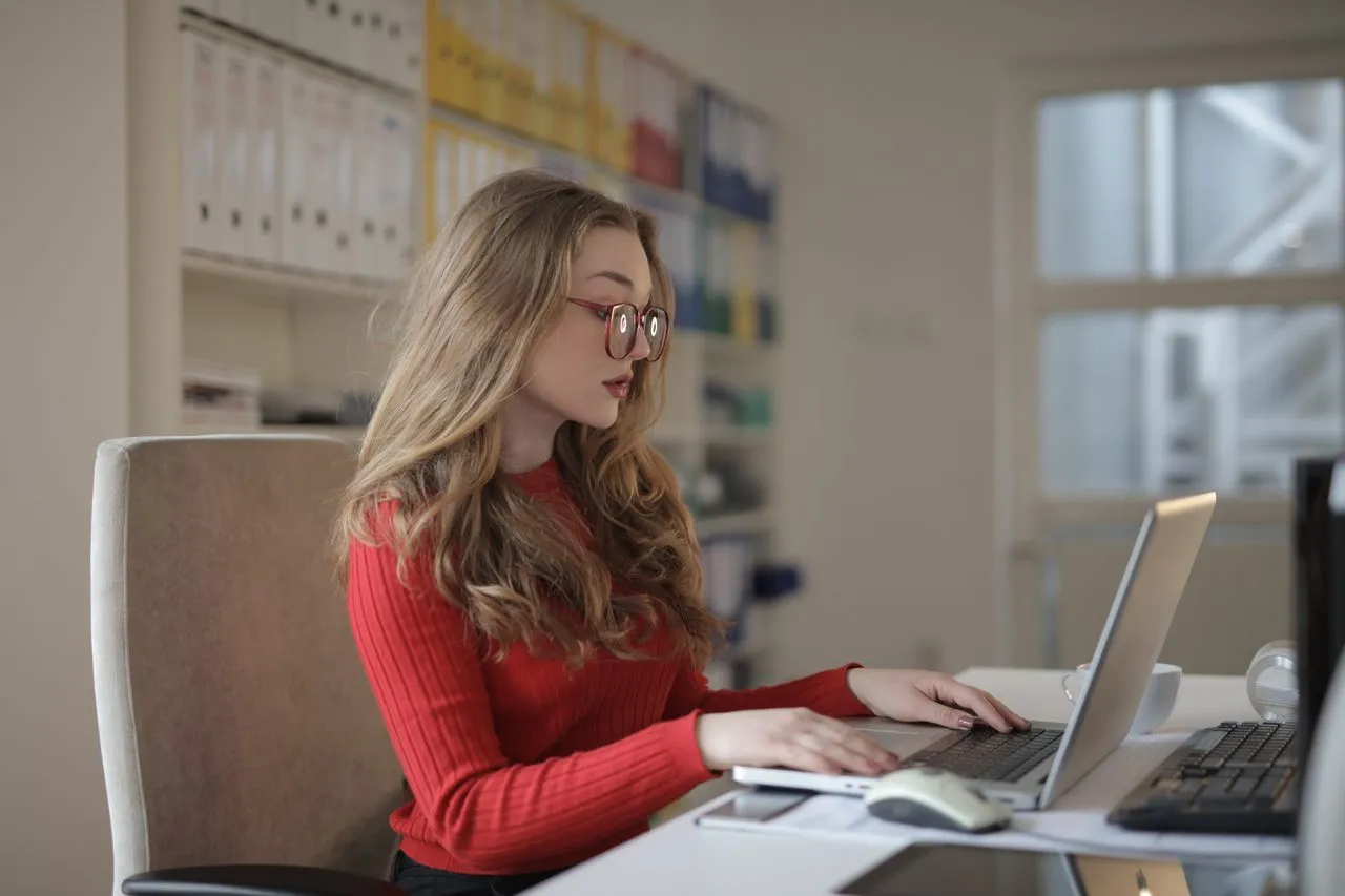 Mulher concentrada, de óculos e blusa vermelha, respondendo em um laptop em um escritório, com prateleiras de pastas coloridas ao fundo