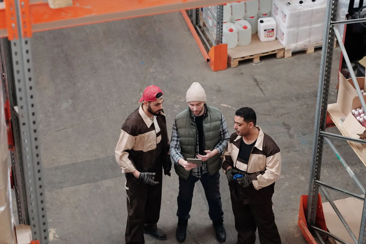 Vista de cima de três trabalhadores de uniforme conversando em pé no corredor de um galpão, um deles mostrando um tablet aos outros dois