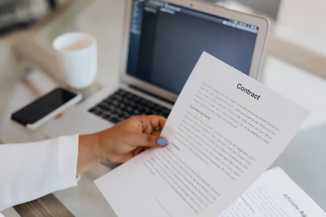Pessoa segurando um documento impresso ao lado de um laptop sobre uma mesa de trabalho.