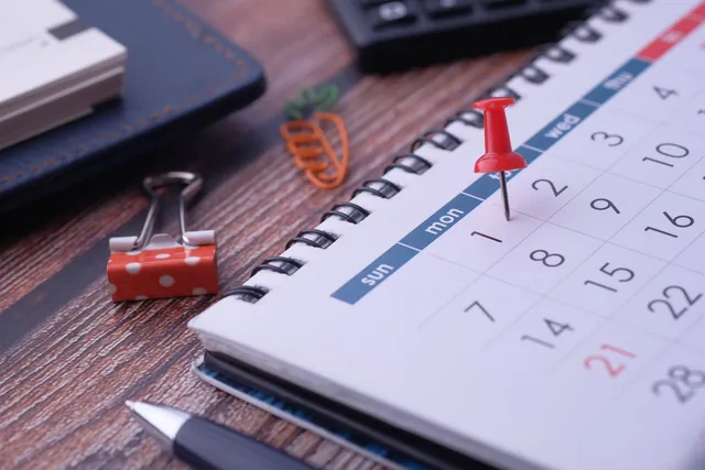 Calendário de mesa com um alfinete vermelho marcando uma data, ao lado de caneta e caderno sobre uma mesa de madeira