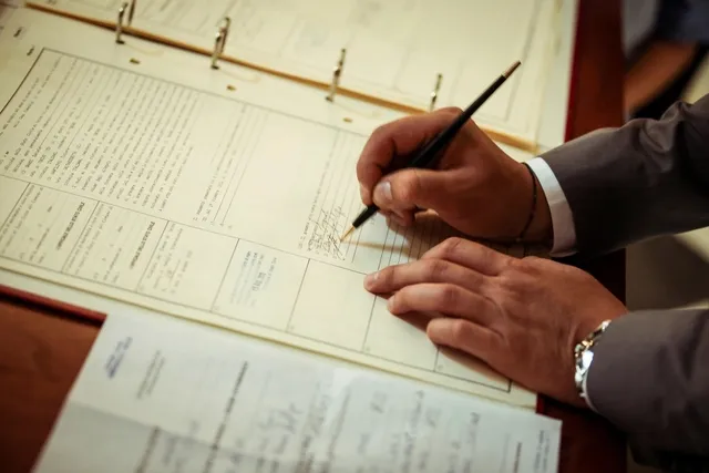 Mãos de uma pessoa de terno preenchendo e assinando um documento formal sobre a mesa de trabalho.